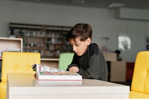 Free A boy in a knitted sweater reads a book attentively in a library. Stock Photo