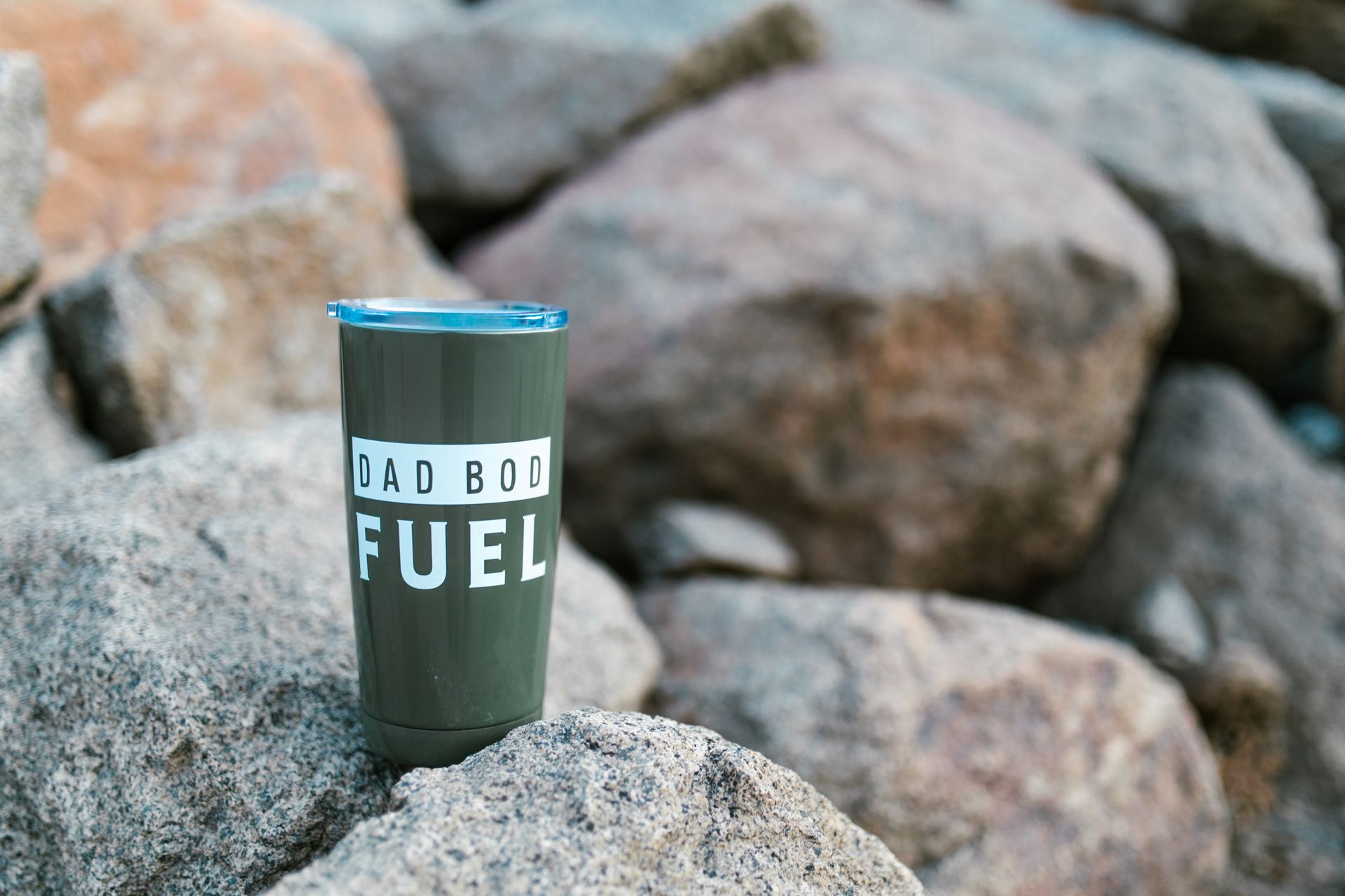 Green tumbler with 'Dad Bod Fuel' text surrounded by rocks outdoors.