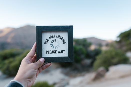 A hand holds a picture frame with 'Dad Joke Loading' text in a natural setting. — photo by RDNE Stock project