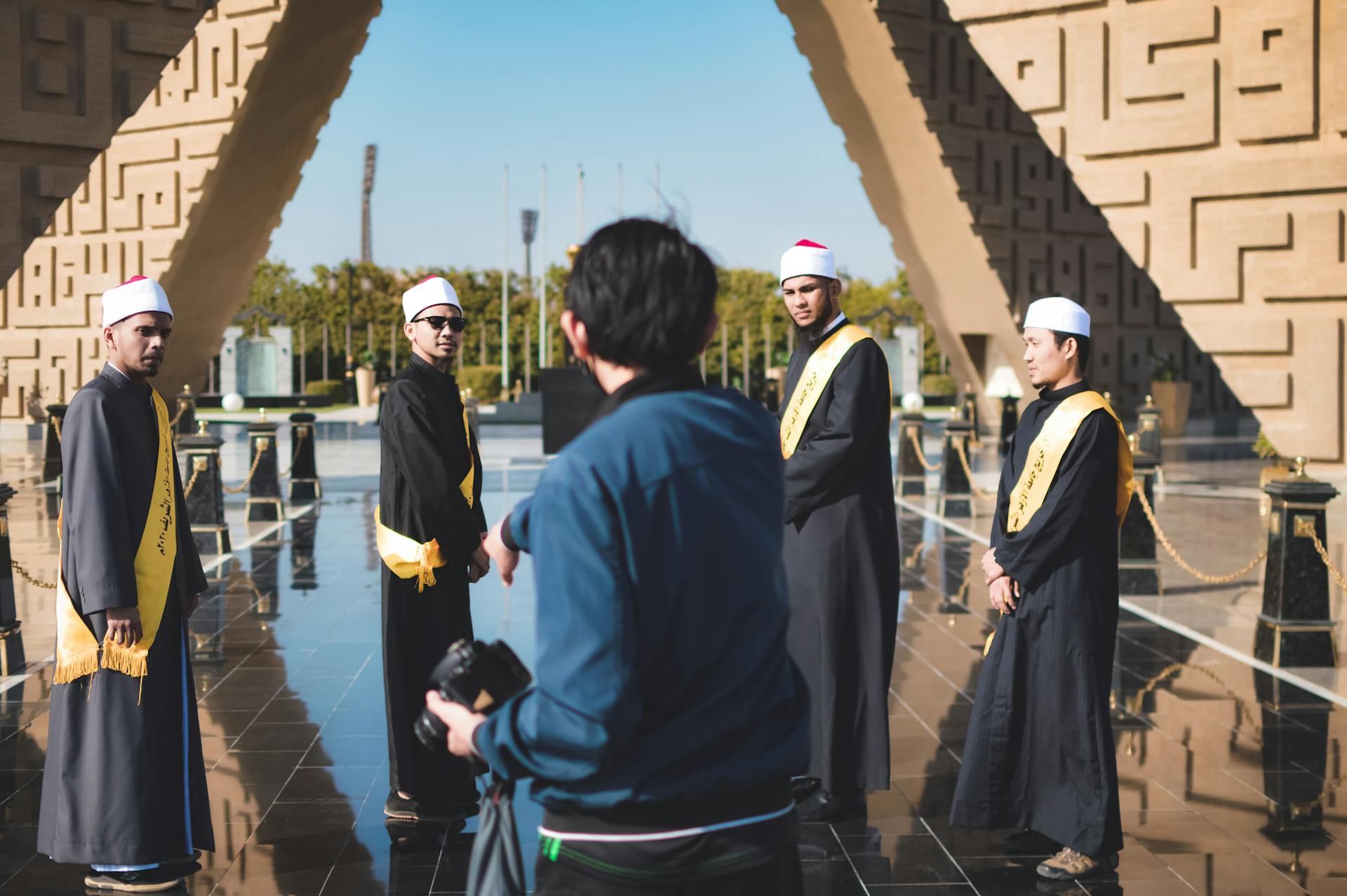 Men in traditional robes stand outdoors during a ceremony under a modern architectural structure.