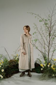 A stylish woman in a trench coat poses gracefully surrounded by spring blooms and minimalistic decor.