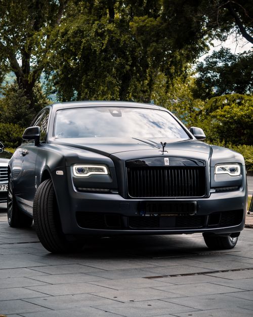 Free A luxury black car parked on a concrete pavement surrounded by green foliage, symbolizing elegance and sophistication. Stock Photo