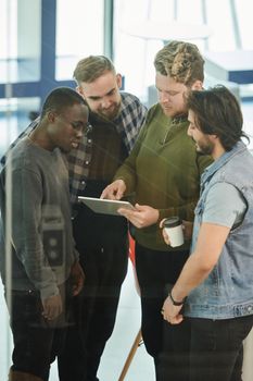 A diverse group of men gathers indoors, discussing content on a digital tablet.