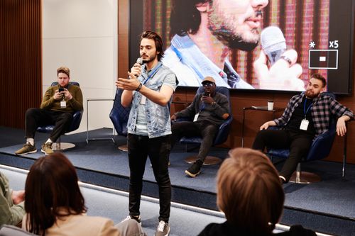 Free Speaker holds microphone, engaging audience at an indoor business conference with a panel in the background. Stock Photo
