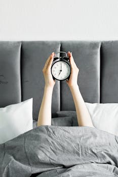 A close-up of hands holding an alarm clock above a cozy bed, signaling morning wakeup.