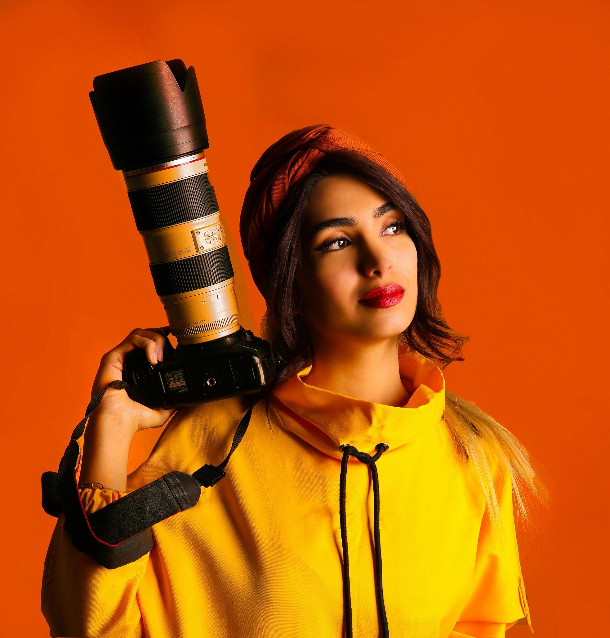 Stylish woman in yellow jacket holds DSLR camera against vibrant orange background.