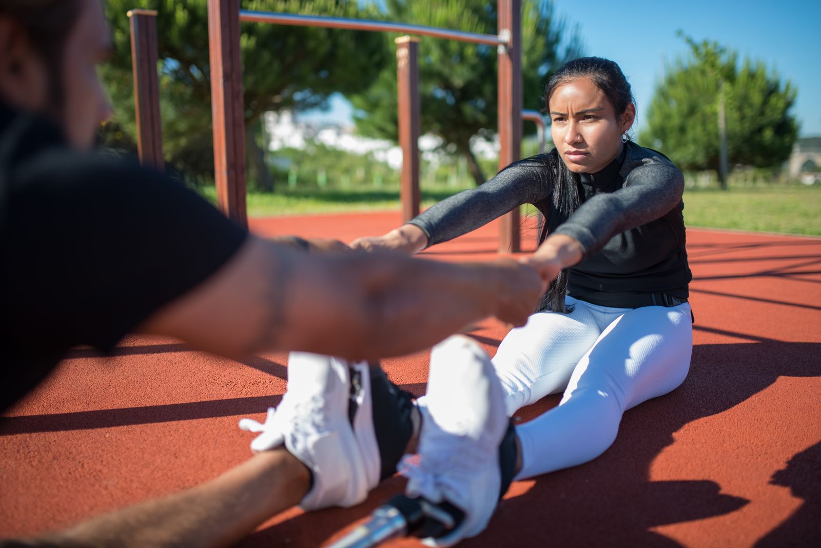 Woman in white leggings stretching on a red outdoor track, holding a partner's hands in sunlight
