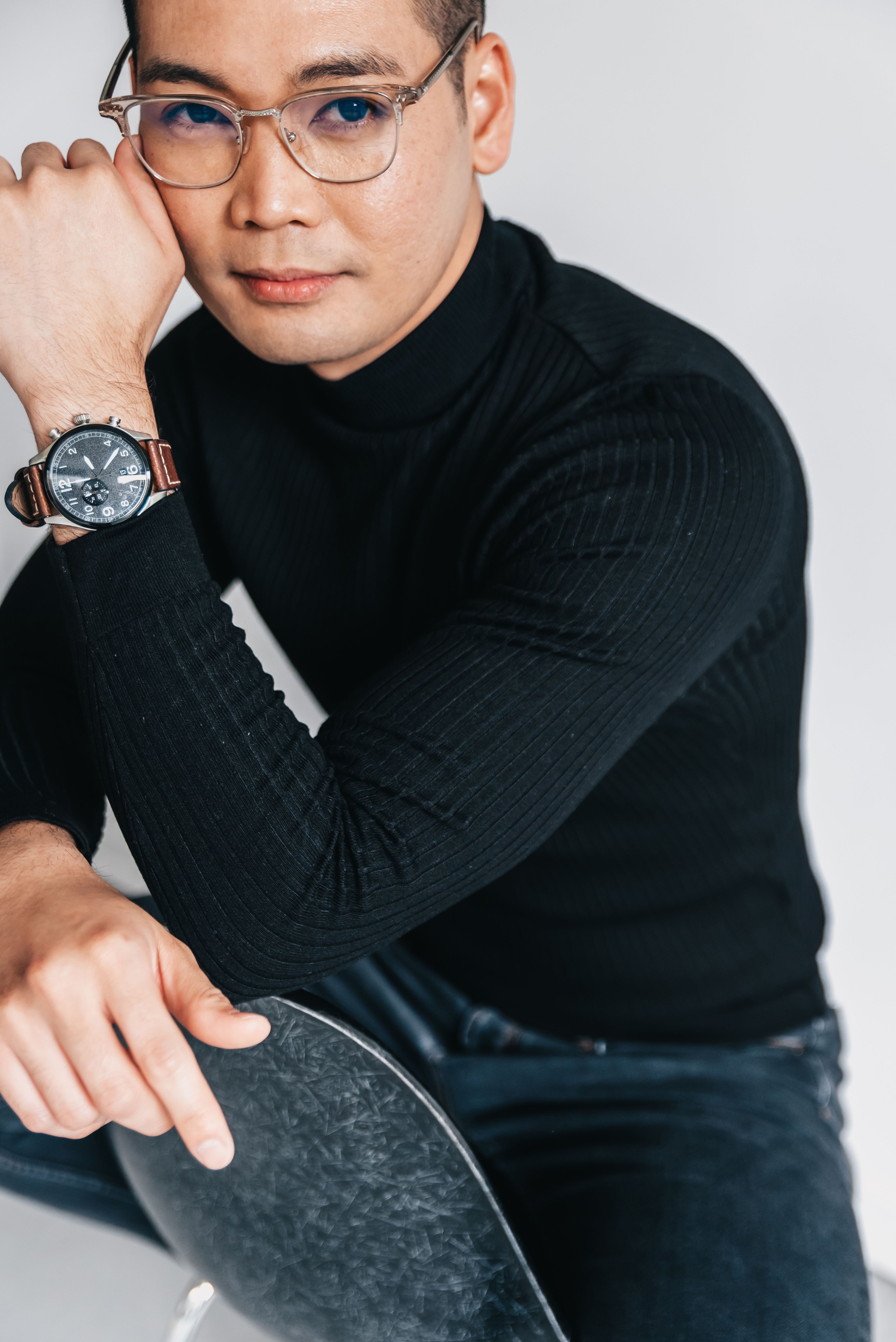 Casual portrait of a young man with eyeglasses and wristwatch sitting indoors against a gray background.