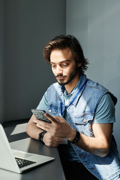 Free Man in denim vest using smartphone and laptop at a desk indoors with sunlight. Stock Photo