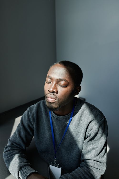 Free African American man sitting peacefully indoors, eyes closed, in sunlight, wearing gray sweater and lanyard. Stock Photo