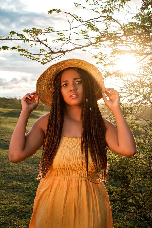 Free A woman in a yellow dress poses outdoors at sunset, highlighting her braided hairstyle and fashion sense. Stock Photo