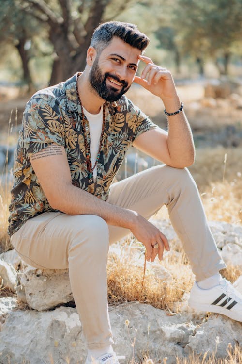 Photo of a Man with a Beard Sitting on a Rock While Looking at the Camera