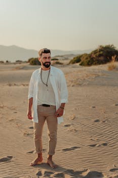 A bearded man in casual attire stands confidently on sunny desert sands.