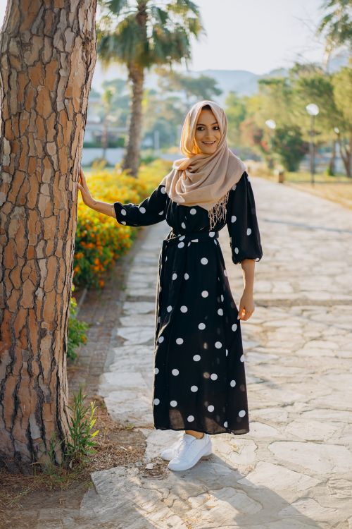 Free Young woman in a polka dot dress smiling and posing by a tree in a sunlit park. Stock Photo
