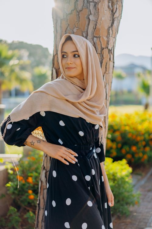 Free Woman in a black and white polka dot dress with a beige hijab standing by a tree outdoors. Stock Photo