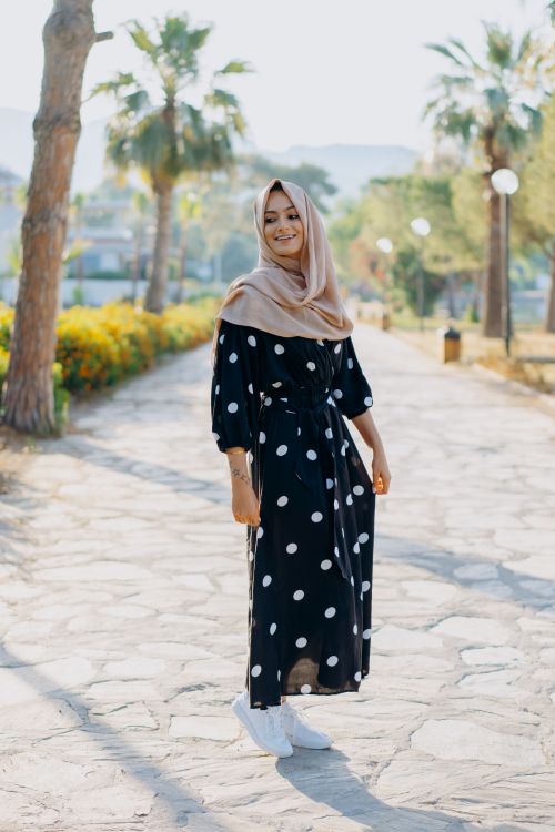 Free A stylish woman in a polka dot dress and hijab smiling while standing on a paved path in a sunny park. Stock Photo
