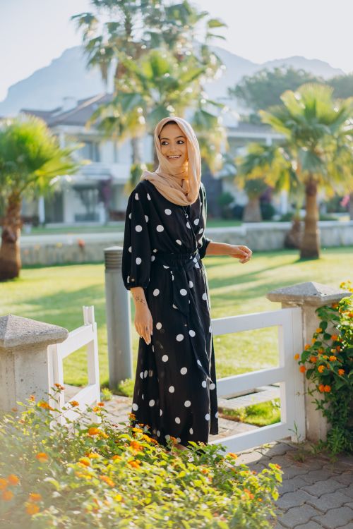 Free Smiling woman in a black polka dot dress and hijab enjoying a sunny day in a garden setting. Stock Photo