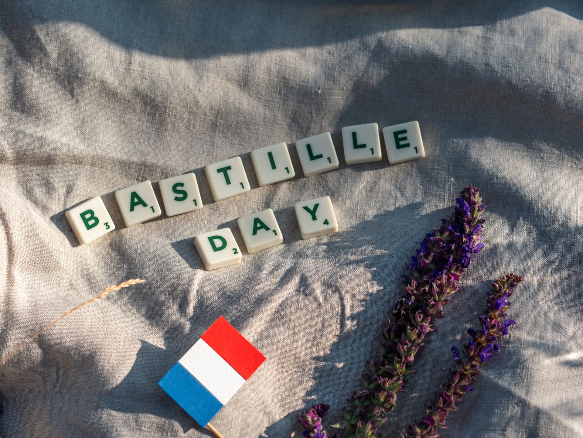 Scrabble tiles spell 'Bastille Day' with French flag and lavender on fabric.