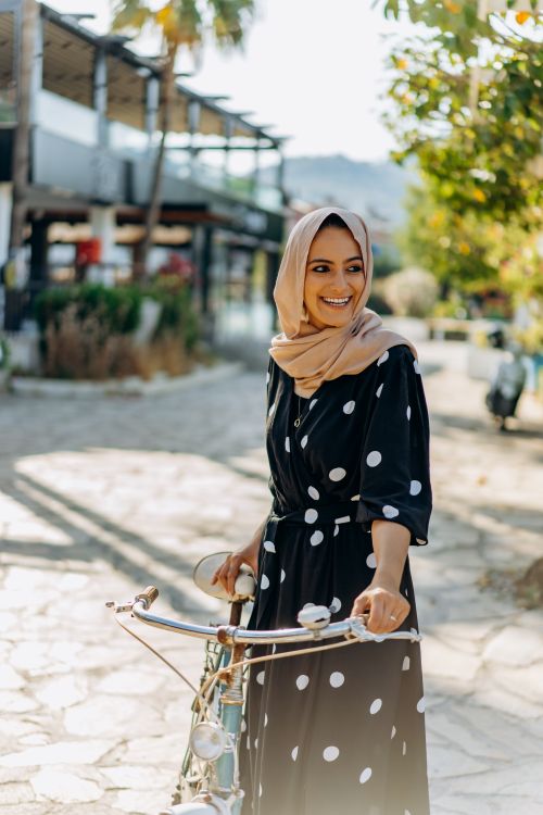 Free Young woman in polka dot dress and hijab smiling while holding a bicycle on a sunlit street. Stock Photo