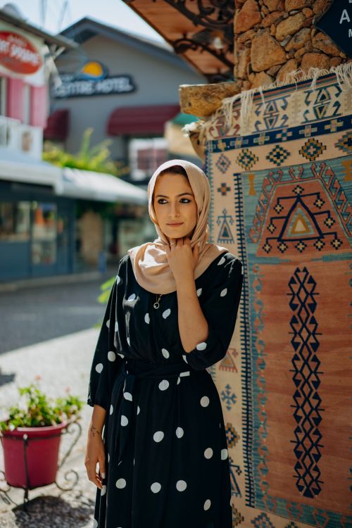 Free Elegant woman in a polka dot dress and hijab posing on a city street beside a decorative rug. Stock Photo