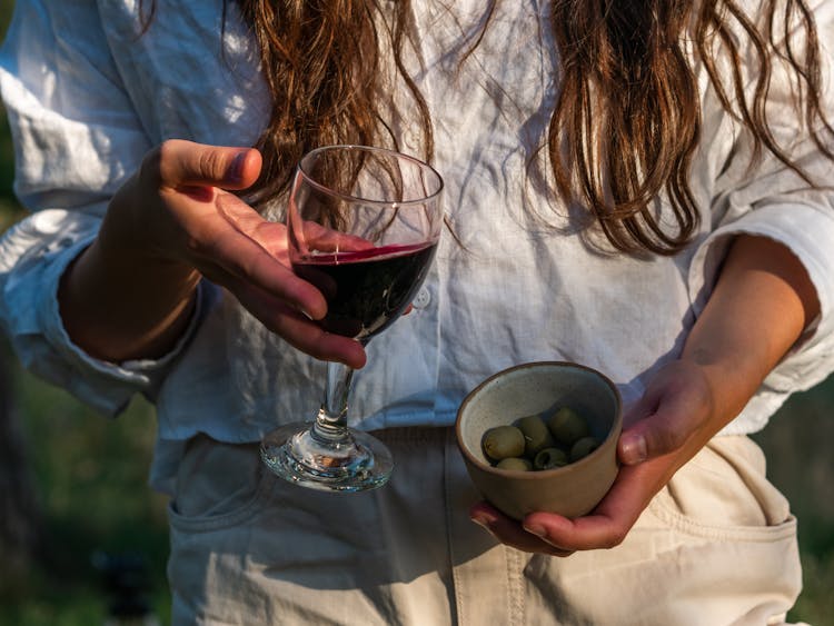 A Woman Holding A Glass Of Wine And A Cup Of Green Olives