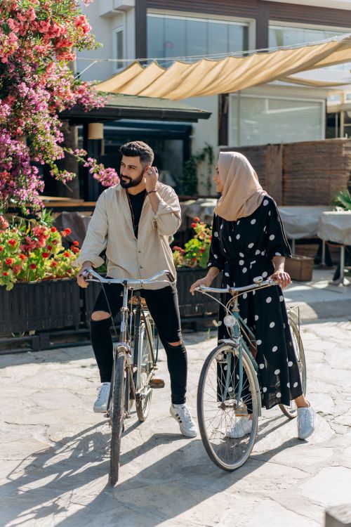 Free A man and woman in stylish attire biking together under sunny skies with floral surroundings. Stock Photo