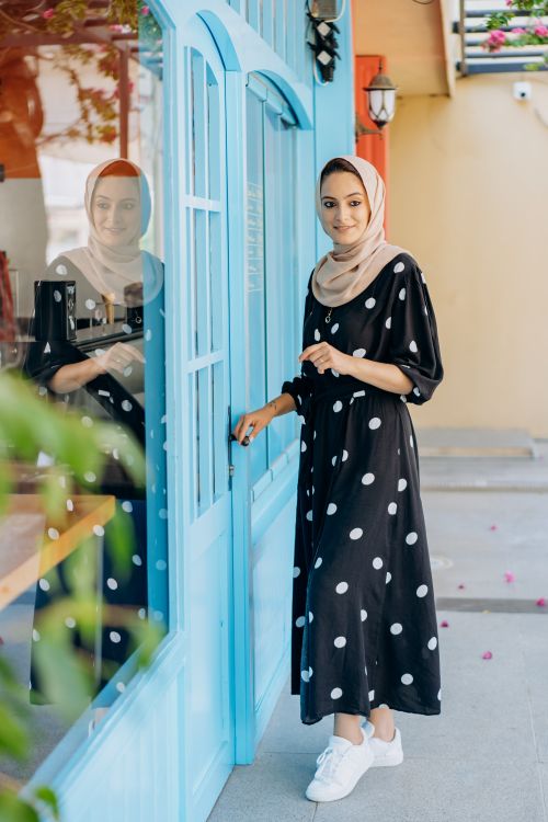 Free Stylish woman wearing a polka dot dress and hijab smiling by a blue glass door outdoors. Stock Photo