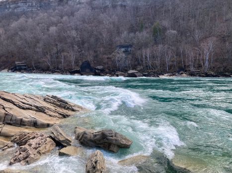 Beautiful view of the Niagara River rapids with rocky foreground and barren trees in winter.