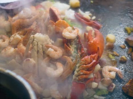 Steaming fresh seafood including shrimp and lobster grilling on a hot plate with vegetables.