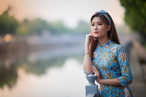 Free Beautiful Asian woman in traditional dress gazing away by a serene riverside with a blurred background. Stock Photo