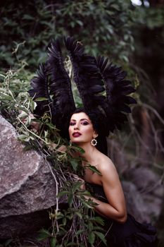 Elegant woman wearing a striking black feather headdress, posing outdoors with lush greenery.