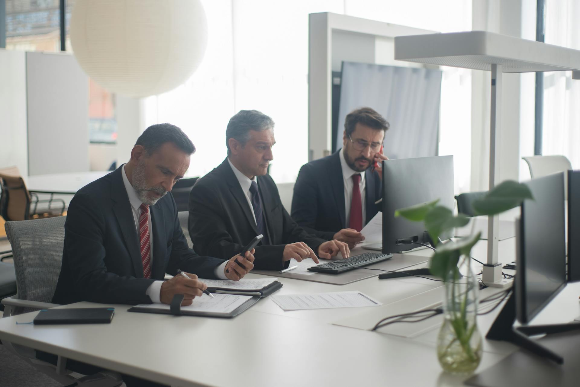 Three men in suits collaborating at a modern office desk with computers and documents.