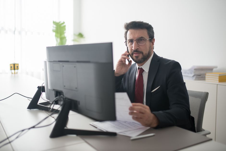 Empresário confiante em um terno preto fazendo uma ligação enquanto revisa documentos em um escritório moderno.