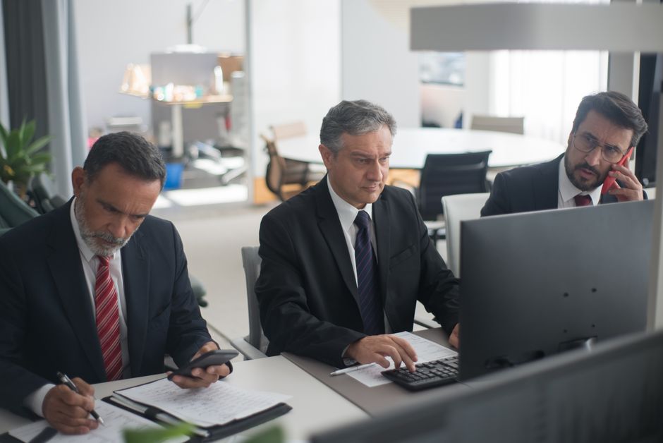 Three businessmen in suits working on computers and documents in a modern office setting. Three businessmen in suits working on computers and documents in a modern office setting.
