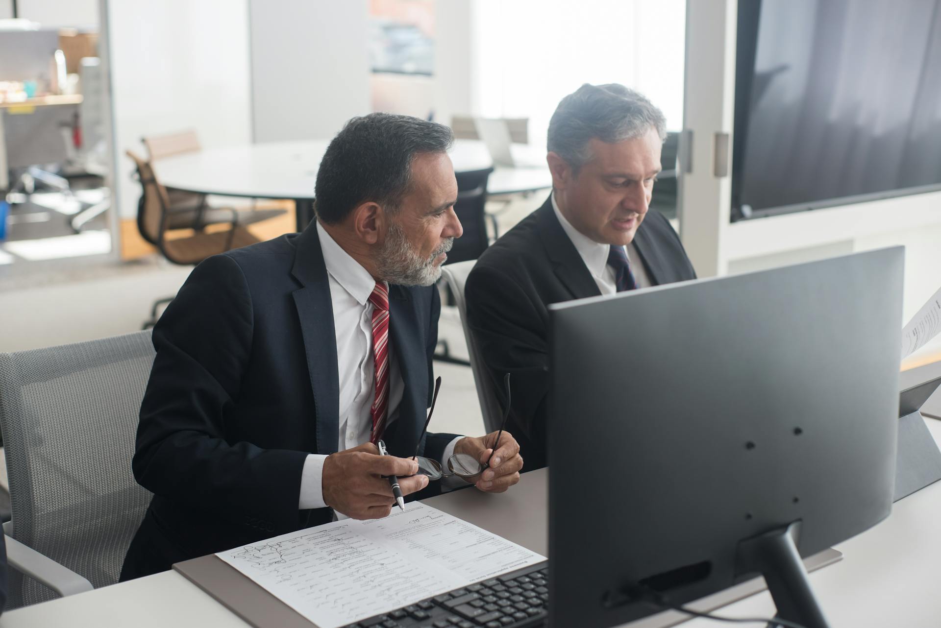 Two male executives in office discussing documents at a computer workstation.