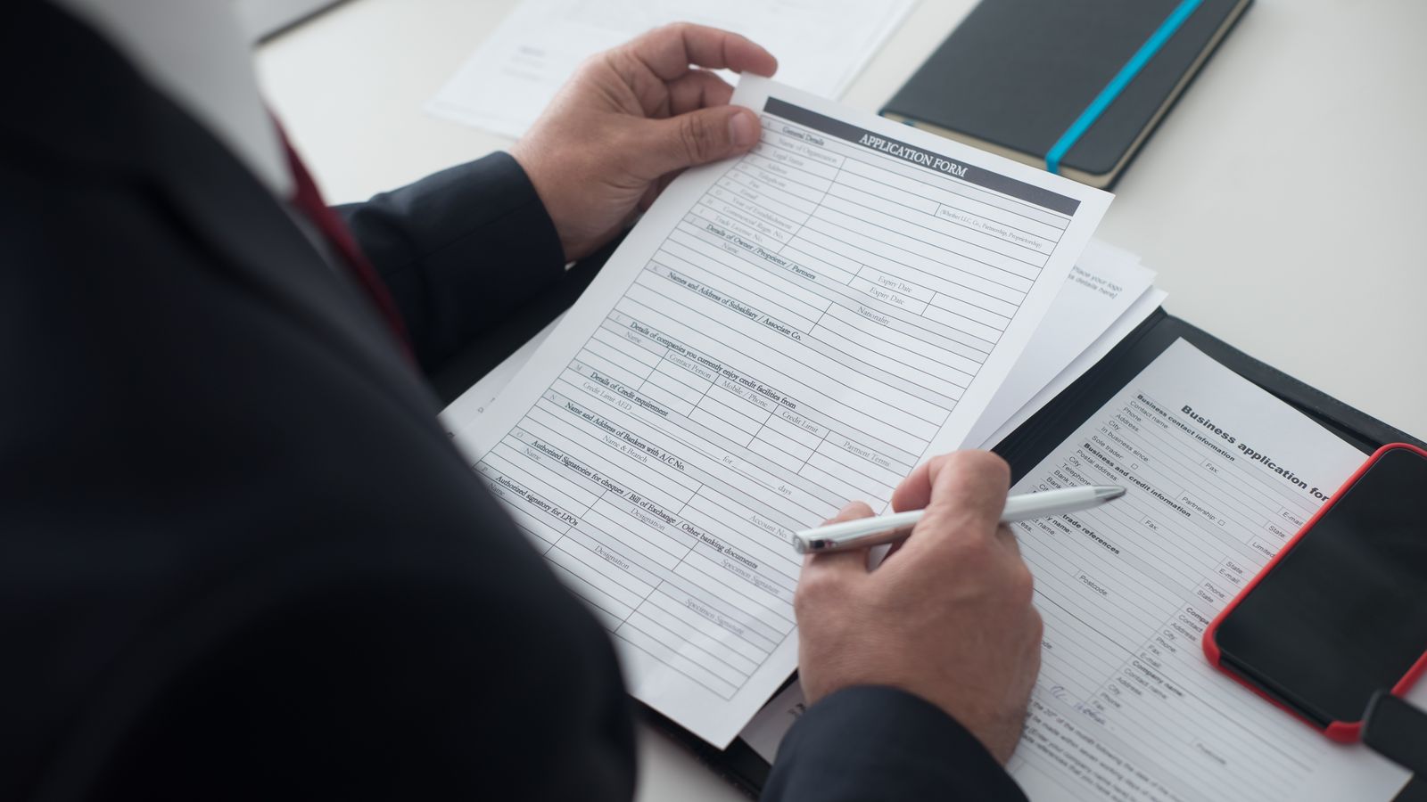 A businessman in a black suit reviews a printed application form with a pen on his desk.
