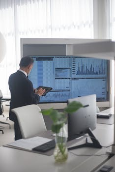 A businessman in a modern office reviews data charts on a large screen with a tablet.
