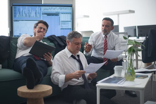 Three businessmen review documents while sitting in an office environment with screens displaying data.