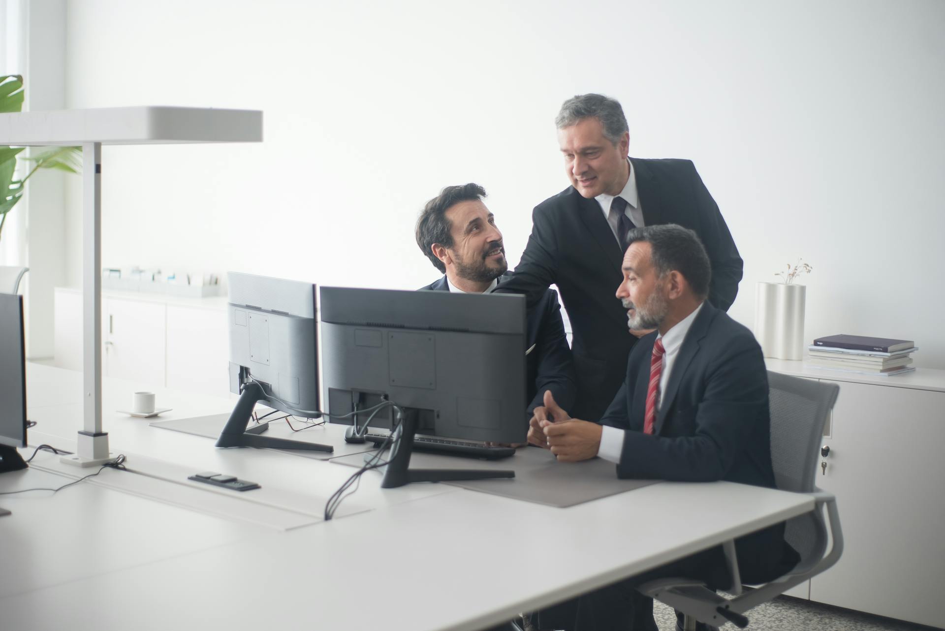 Three business executives in suits collaborating around dual monitors in a modern office setting.