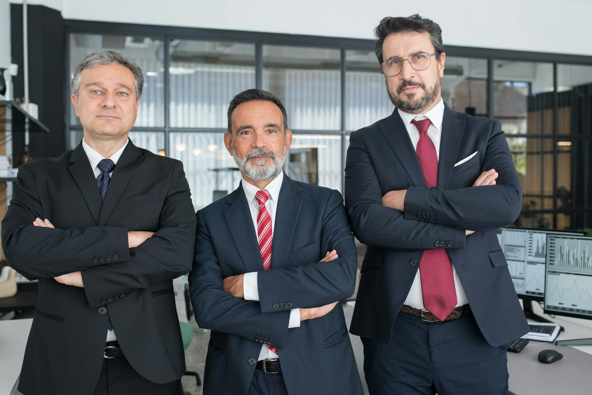 Three senior business executives in suits standing confidently in a modern office environment.