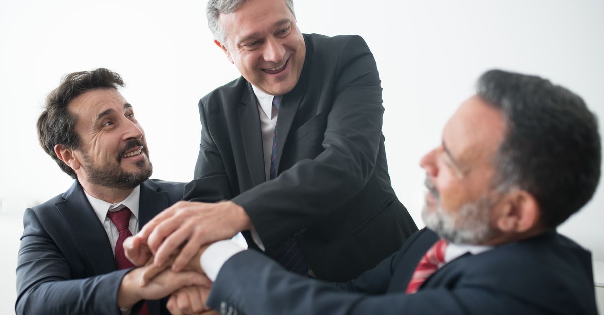 Three businessmen in suits celebrating teamwork success indoors with handshake.