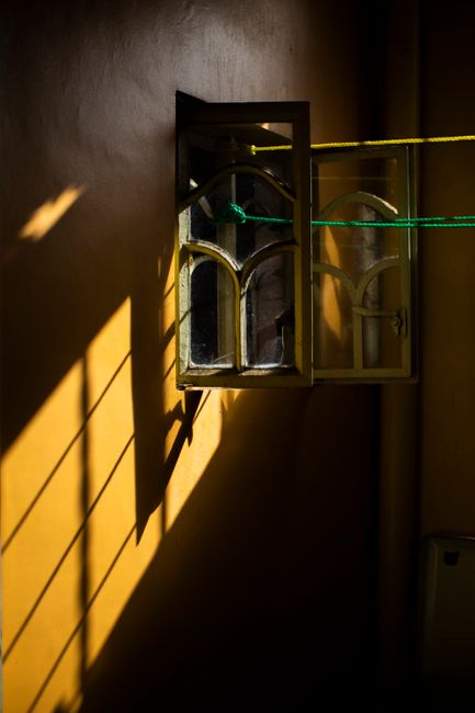 Warm sunlight casts dramatic shadows through an open vintage window against a bright yellow wall.