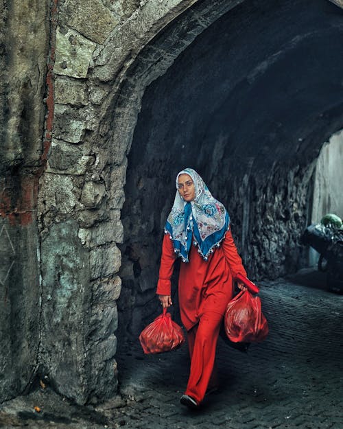 A Woman in Red Long Sleeves and Pants Walking on the Street while Carrying a Plastic Bags