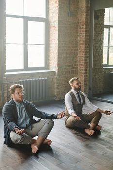 Two men practice meditation indoors, sitting in a peaceful pose with eyes closed under natural light.
