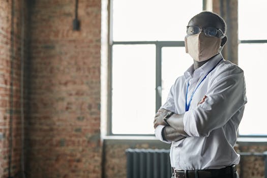 A man with a face mask and crossed arms stands confidently in a brick-walled office.