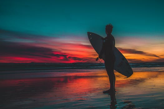 Silhouette of a surfer holding a board on a beach at sunset with colorful sky reflections.