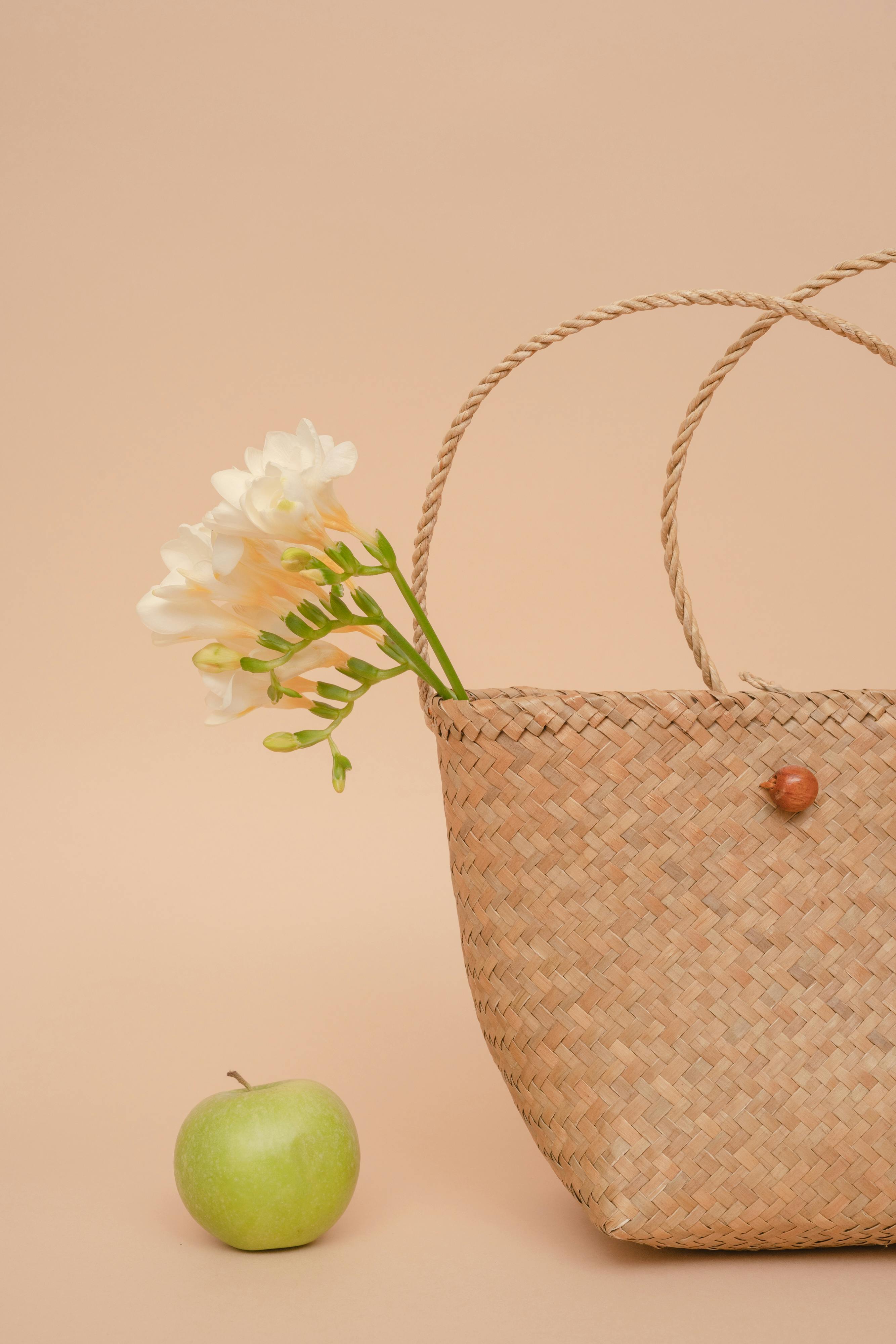 Still life with woven handbag, green apple, and white flowers on beige background.