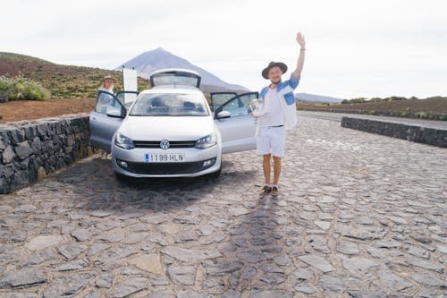 Free Cheerful male in white clothes standing on cobblestone near car and greeting friend while resting with wife in countryside Stock Photo