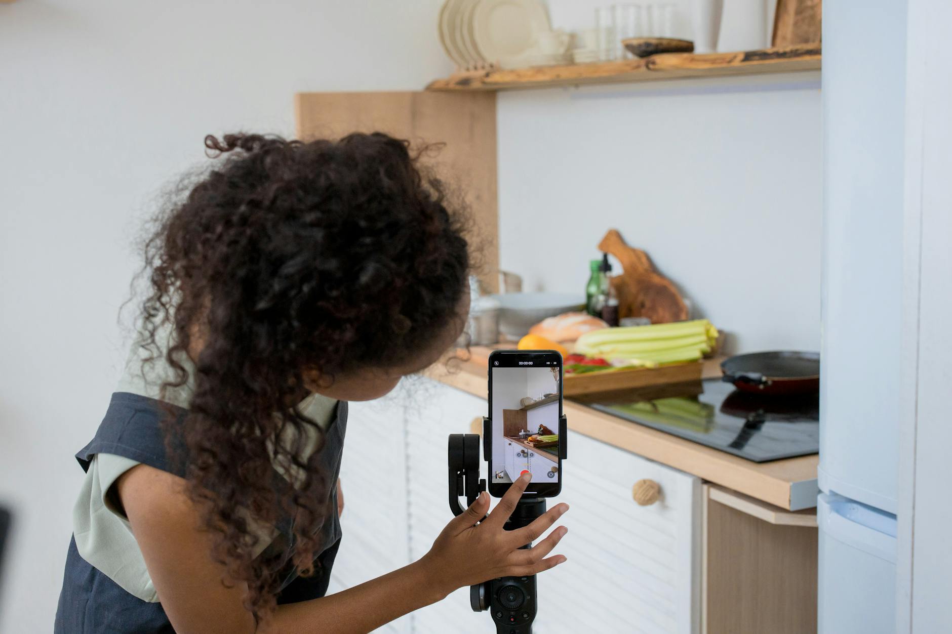 A woman with curly hair films a cooking video using a smartphone stabilizer in a modern kitchen.