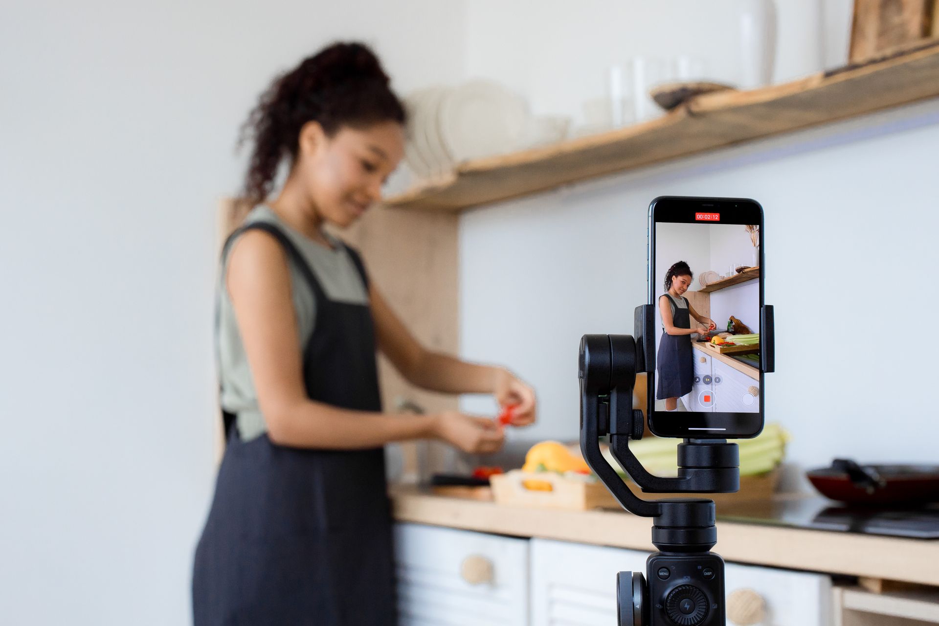 A woman records a cooking tutorial with her smartphone in a modern kitchen setting.